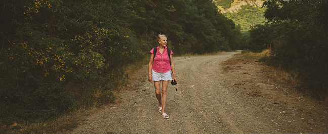 Excited happy senior woman backpacker tourist walking in summer forest road outdoors at sunset time. Old slim lady traveling with photo camera. Active retirement vacation concept. Warm filter