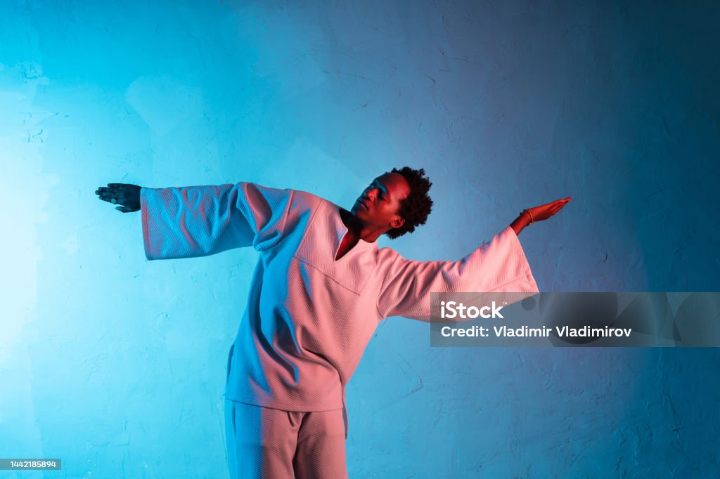 Man dancing with arms held wide An African-American man twisting his body and twisting his arms, wearing modern Japanese style clothing against a gradient blue background 20-24 Years Stock Photo