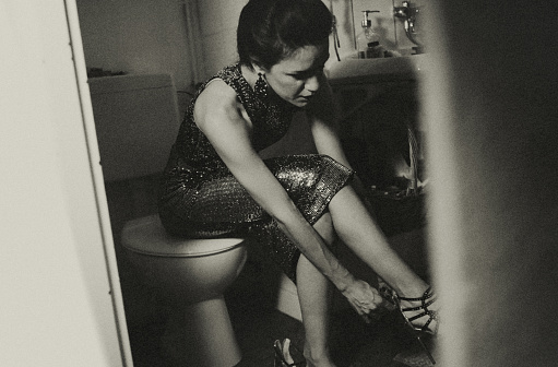 Portrait of a young woman in her bathroom. She is getting ready for a party. She is wearing a sequined cocktail dress and just putting on her high heels.