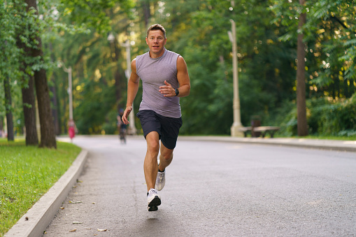 Healthy active man runner running in park. Handsome young male jogger trains cardio going for a run in city park. Lush green foliage of trees, road in the background in summer. He is looks in camera