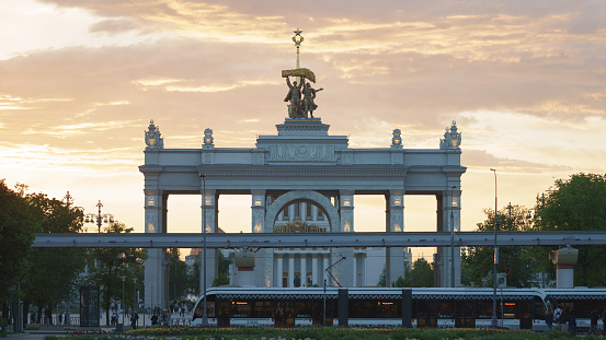 Moscow, Russia - May 23, 2021: Soviet architecture of the main entrance into the park VDNkH in spring day. A lot of people. Tractor driver and collective farmer statues. Tram drives on foreground