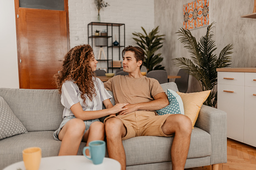 Beautiful happy young couple sitting on the sofa in the living room talking and drinking coffee.