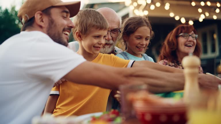 Happy Senior Grandfather Talking and Having Fun with His Grandchildren, Holding Them on Lap at a Outdoors Dinner with Food and Drinks. Adults at a Garden Party Together with Kids.