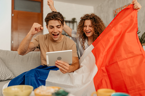 Beautiful young happy couple, holding a French flag, rooting for their team while watching the game on a digital tablet. Scoring in the game.