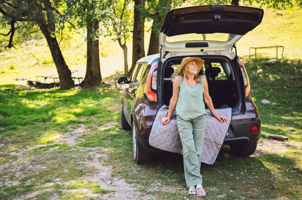 Happy senior woman in straw hat sitting in car trunk or boot outdoors in camper sunny summer park, smiling enjoying journey. Retired people activity and road trip concept. feliz mujer mayor con sombrero de paja sentada en el maletero del automóvil o en el maletero al aire libre en el soleado parque de verano camper, sonriendo disfrutando del viaje. actividad para jubilados y concepto de viaje por carretera