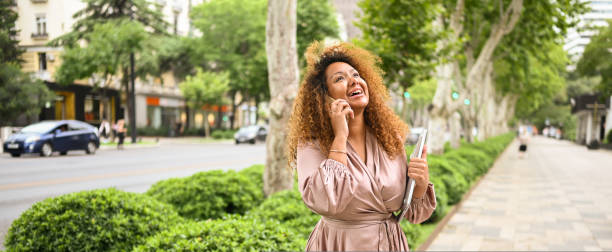 Attractive happy stylish plus size African American woman student freelancer afro hair with laptop computer outside. Young female office worker speaks phone in big urban city summer street. Diversity. stock photo