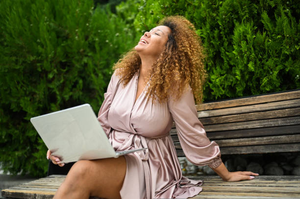 Attractive happy stylish plus size African American woman student freelancer afro hair studying online working on laptop computer outside at summer park. Diversity. Remote work, distance education. stock photo