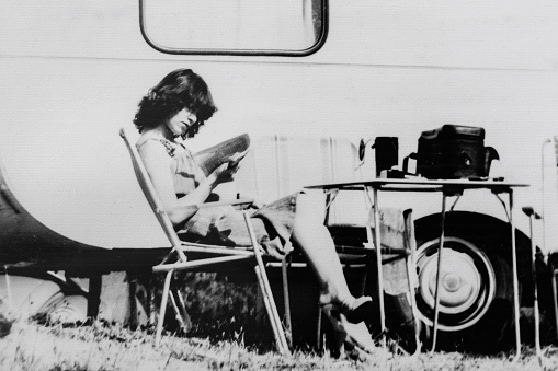 Black and white photo of woman reading a book next to camper