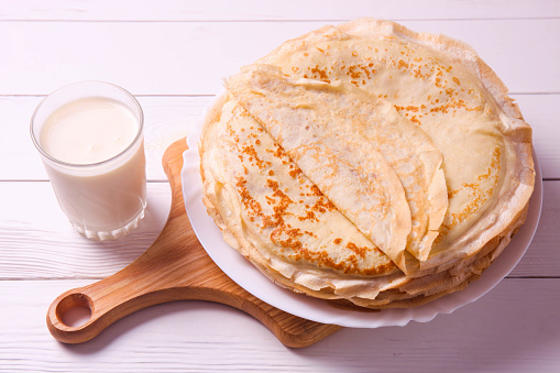 Stack of thin pancakes on plate and glass of milk on white wooden background. Pancake week