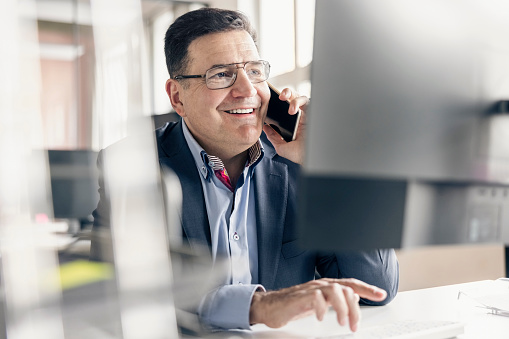 Happy mature businessman talking on phone while working at his desk in office. Smiling mid adult businessman answering a phone call at work.
