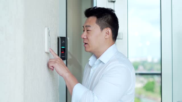 Asian man, calling rings the intercom of the house, pressing the button, waiting for the door to open. Male pushing the button and talking doorbell with camera near