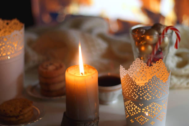 Glass with Christmas ornaments, cup of tea, cookies, candles and knitted blanket by the fireplace. Cozy Christmas at home.