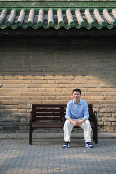 one smiling Asian young man sitting on bench in front of palace wall, looking at camera