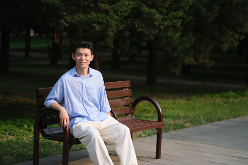 smiling handsome Asian young man, sitting on bench in park on sunny day. Looking at camera