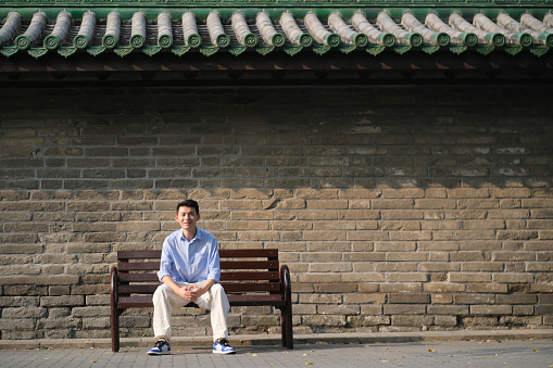 one smiling Asian young man sitting on bench in front of palace wall, looking at camera