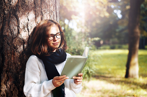 Young adult brunette woman in glasses and a scarf reading a book in the park.