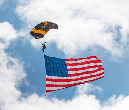 Farmingdale, New York, USA - 26 May 2022: A United States Army paratrooper on the Golden Nights Team opens up the airshow parachuting down to the ground with a very large American Flag.