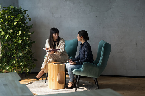 Two friendly women colleagues seated in armchairs enjoy pleasant talk, share information at informal meeting in modern lobby of office. Indian and Caucasian businesswomen conversation during workflow