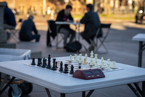Two teenage boys are playing chess match against one man in city park. They are playing street cheese with huge table board and figurines.