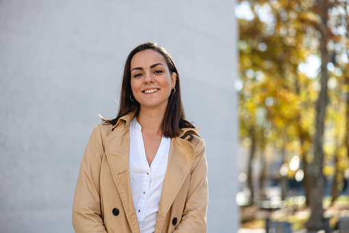 Waist up front shot with blurred background of a young and beautiful woman with professional make-up and long brown hair smiling and standing in front of a white building and a park while wearing a beige trench coat on a sunny autumn day.