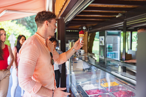 Young attractive caucasian man holding a two flavor ice-cream cone in front of the counter of an ice-cream shop. There are some multi-racial people behind him waiting and chatting.