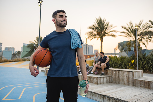 Young Adult Male Holding a A Basket Ball And A Water Bottle Standing Looking And Smiling Away From The Camera In Dubai.