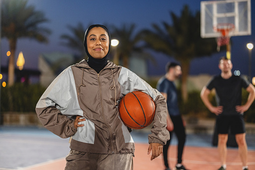 Portrait Young Adult Female Athlete Holding A Basket Ball With One Hand And The Other Hand Is On Her Hip. She Is Smiling With Close Mouth Looking At The Camera Outdoors At Night In Dubai.