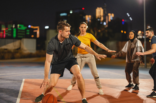 Multi Ethnic Young Adults Playing Basketball Outdoors At Night In Dubai.