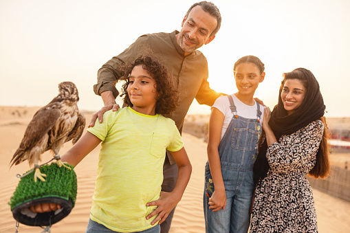 Portrait of a  happy Middle Eastern family in the dune. The youngest daughter is proudly having a falcon on her hand in Dubai.