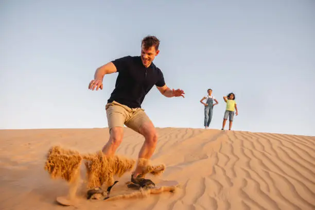 Young adult male with sport clothes on vacation trying sandboarding in Dubai. There are young teenage girls in the back waiting for their turn.