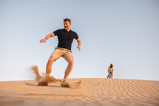 White male in motion. He is sandboarding down the dune in Dubai. He is  standing on the board excited and happy.