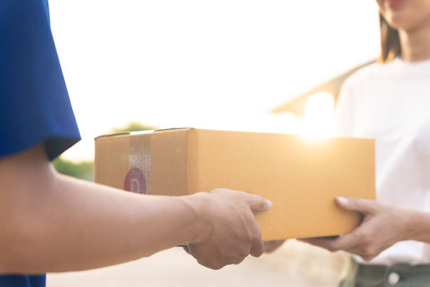 istock Close up delivery man hands wearing blue uniform send a cardboard box to customer in front of the customer's house with sunset flare background. 1442262353
