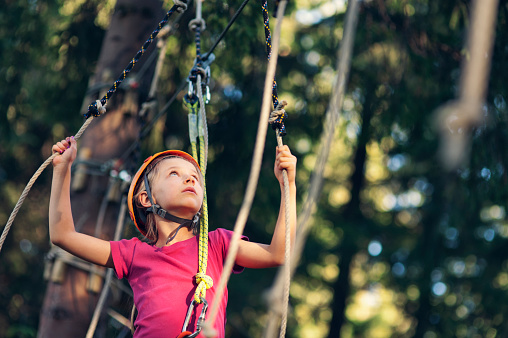 Focused little girl wearing a helmet is walking and climbing in high ropes course in adventure park. \nSunny summer day evening. \nNikon D700
