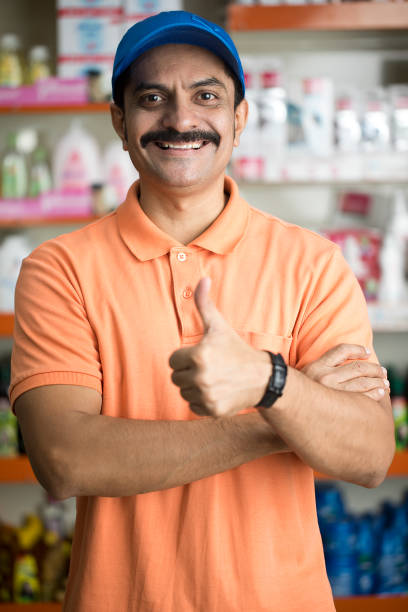 Happy owner showing thumbs up in store Portrait of smiling Indian male owner wearing cap showing thumbs up while standing in supermarket convenience store stock pictures, royalty-free photos & images