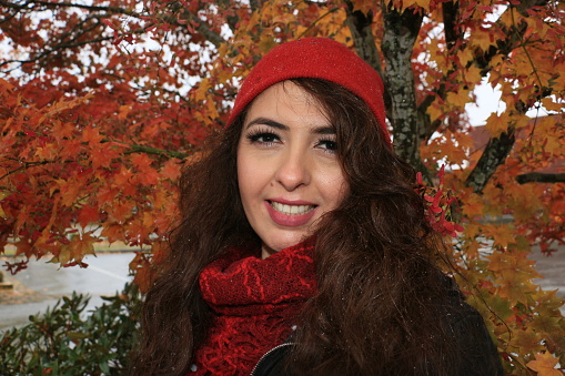 A Mexican woman in front of a tree in October with its leaves in full color. She is wearing a red toque and scarf to keep warm and a brown fully unbuttoned jacket. She is also wearing makeup.