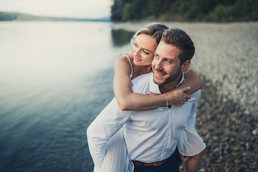 Smiling husband carrying his wife piggyback, standing in shallow water of river on late summer afternoon, waist up, background blurred