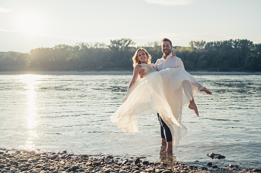 Beautiful wedding couple in shallow water of river in backlight on sunny summer afternoon, groom carrying bride, both smiling at camera