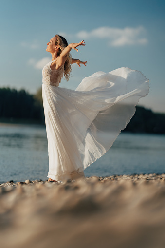 Beautiful woman in white wedding dress standing alone at riverbank with arms outstretched