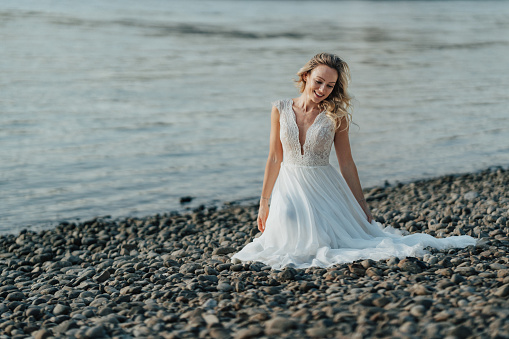 Young woman in white wedding dress sitting alone on beach with stones close to river