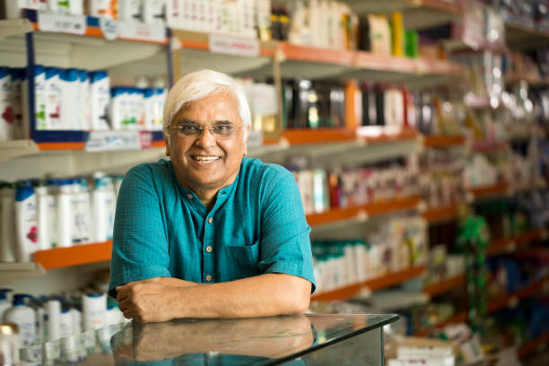 Portrait of smiling Indian senior male manager leaning on checkout counter while standing against products at supermarket