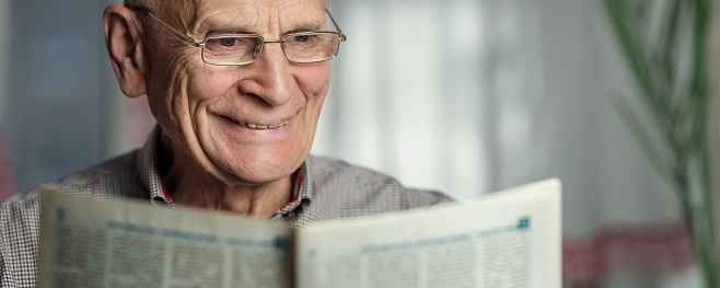 Elder man wearing eyeglasses reading newspaper and smiling. Retirement leisure and spending time concept.