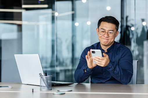 Dissatisfied asian businessman got bad news, man reading on smartphone sitting inside modern office with laptop, disappointed boss with phone upset.