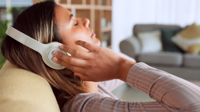 Woman, headphones and relax listening to music on living room sofa with eyes closed at home. Female enjoying calm audio track with wireless headset relaxing in comfort on couch at the house indoors