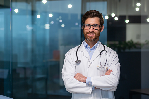 Portrait of mature doctor with beard, man in white medical coat smiling and looking at camera with crossed arms working inside modern clinic.