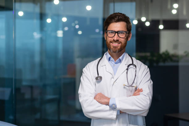 portrait of mature doctor with beard, man in white medical coat smiling and looking at camera with crossed arms working inside modern clinic - médico geral imagens e fotografias de stock