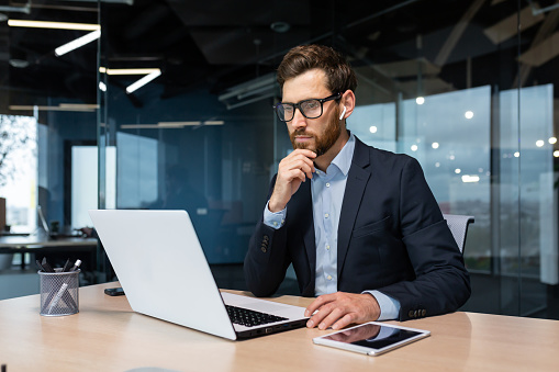 Senior businessman in glasses and beard is thinking about a decision, man is working inside the office and using laptop at work, mature investor in a business suit is sitting at the table