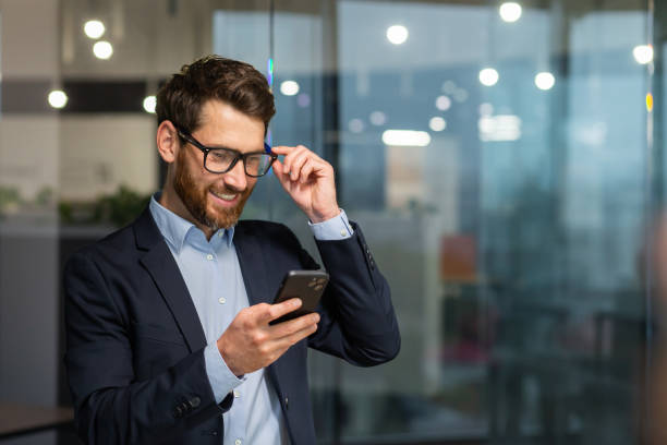 l'investitore finanziario di successo lavora all'interno dell'ufficio al lavoro, l'uomo d'affari in giacca e cravatta usa il telefono vicino alla finestra, l'uomo sorride e legge buone notizie online dallo smartphone - businessman telephone men business foto e immagini stock