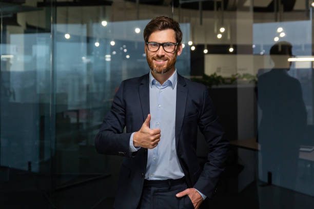 portrait d’un patron d’homme d’affaires mûr à succès, d’un gestionnaire en lunettes de costume d’affaires et barbe regardant la caméra et souriant debout près de la fenêtre, montrant le pouce levé, signe de succès et d’atteinte des obje - working smiling equipment car photos et images de collection