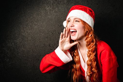A happy young girl in a Santa Claus costume smiles on a black background. Portrait of a red-haired woman in a red cap screaming on a dark background. The concept of Christmas and New Year.
