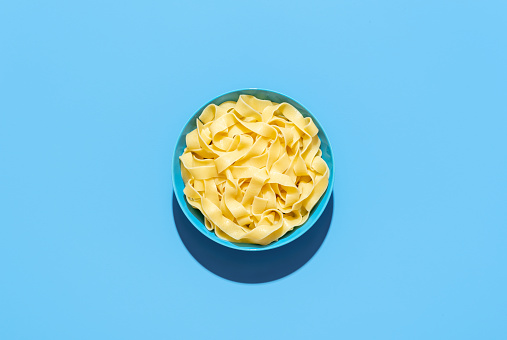 Top view with a bowl with cooked pappardelle pasta, minimalist on a blue table. Simple plain noodles in bright light on a vibrant colored background